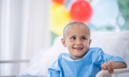 ילד צעיר מחייך במיטת בית חולים עם בלוני צבע ברקע, במסגרת תוכן על קצבת ילד נכה החולה בסרטן מטעם אופקים מימוש זכויות.