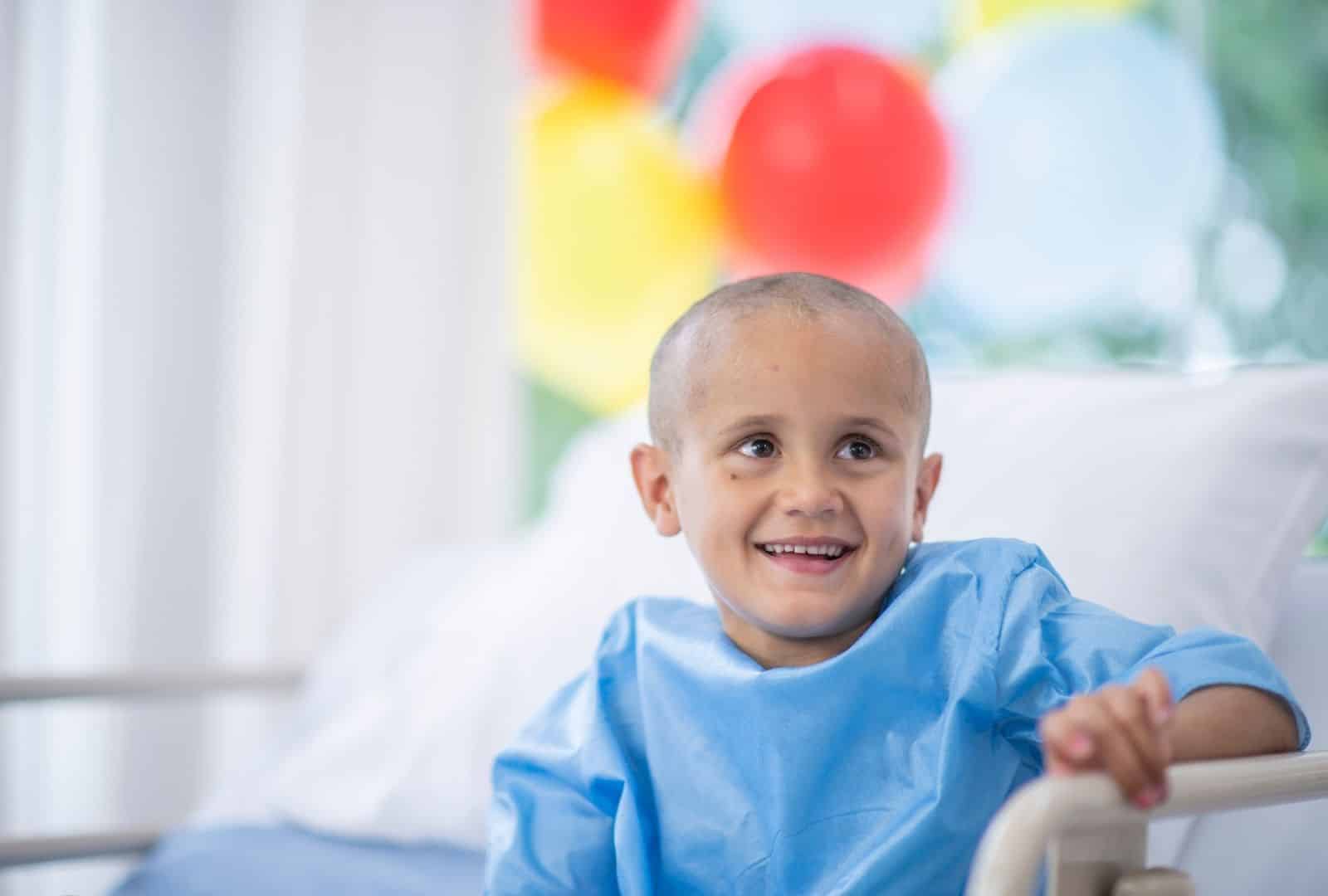ילד צעיר מחייך במיטת בית חולים עם בלוני צבע ברקע, במסגרת תוכן על קצבת ילד נכה החולה בסרטן מטעם אופקים מימוש זכויות.