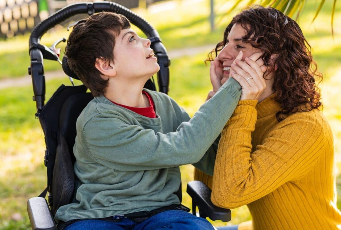 אמא מחבקת ומחייכת אל פניה של בנה הקטן בכיסא גלגלים, רגע אינטימי של חום ואהבה בפארק שטוף שמש, עם דשא ירוק ועצי דקל ברקע – סמל להתמודדות משפחתית עם אתגרי נכות.