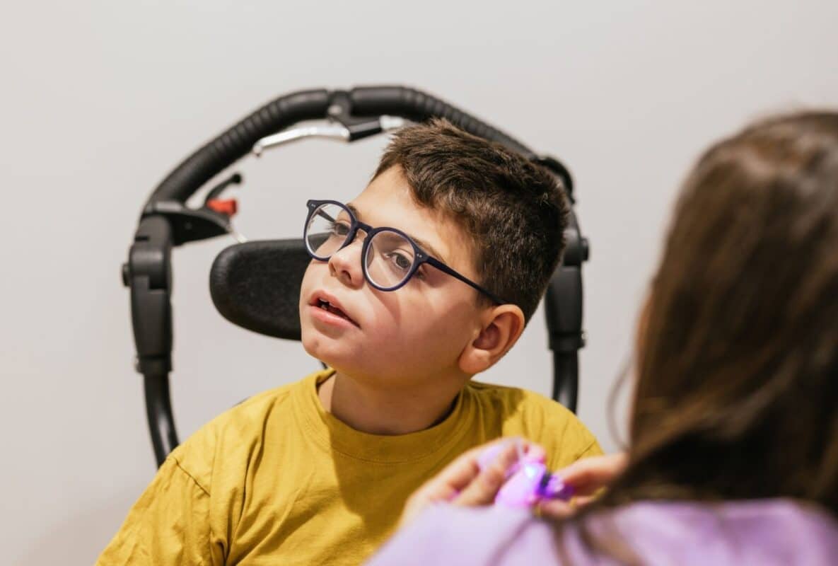 ילד צעיר בכיסא גלגלים עם משקפיים כחולים, מחייך ומסתכל באהבה לאישה (אמו?) המחזיקה בידו צעצוע ורוד זוהר, בחדר בהיר ומזמין – סמל לאהבה, תמיכה ומאבק יומיומי של משפחות עם ילדים בעלי מוגבלויות.